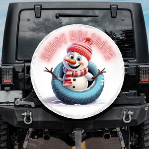 Könnte beinhalten: Eine weiße Reserveradabdeckung mit einem Cartoon-Schneemann, der einen rot-weißen Schal und einen rosa Hut trägt. Die Radabdeckung hat die Worte "Happy Holidays" in pinken Neonbuchstaben.