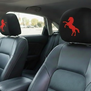 May include: Black car seat headrests with a red horse logo. The headrests are installed in a car with black leather seats. The horse logo is a stylized silhouette of a rearing horse.