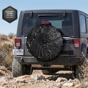 May include: A black spare tire cover with a topographic map design is mounted on the back of a gray Jeep. The cover has a textured, layered appearance. The Jeep is parked on a rocky, dirt path. A badge on the left reads "HEAVY DUTY OPTIONS - 1000+ SALES - ORIGINAL DESIGNS."