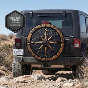 May include: A black Jeep with a spare tire cover featuring a compass design. The compass rose is gold with black accents, and the tire cover has a weathered, textured appearance. The license plate reads "JEEPOTO".