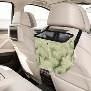 May include: A green and beige patterned trash can with a black rim hangs from the headrest of a car seat. The trash can has a black strap with a buckle.