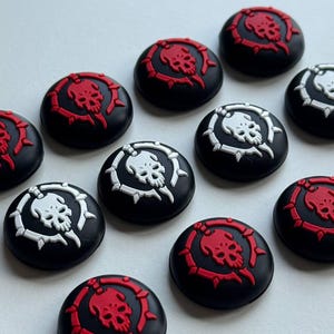 May include: A collection of black, circular buttons with a skull and bone design. The skull and bone design is featured in red and white. The buttons are arranged on a white surface.