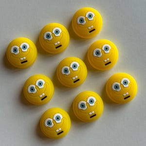 May include: A collection of ten yellow, circular decorative buttons featuring the face of a cartoon character with blue eyes, a smiling mouth, and two square teeth. The buttons are arranged on a white surface.
