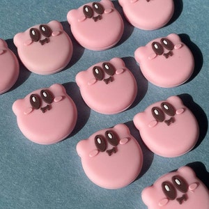 May include: A collection of pink, circular embellishments featuring a cartoon character design. Each charm has a light pink base with brown eyes, a small mouth, and tiny arms. The charms are arranged on a light blue surface.