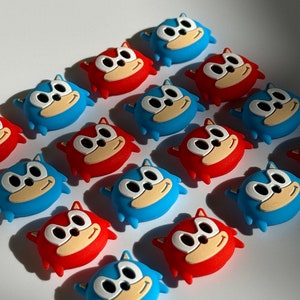 May include: A collection of 14 blue and red cartoon character buttons. The characters have white eyes and a white smile. The buttons are arranged in a staggered pattern.
