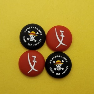 May include: Four round black and red buttons with different designs. Two buttons feature a black and white skull and crossbones with the text "MONKEY.D.LUFFY STRAW HAT PIRATES". The other two buttons feature a red background with a white stylized sword design.