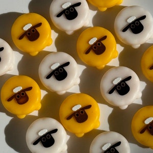 May include: Close-up of several sheep-shaped buttons. The buttons are either white or yellow, with a black face, brown ears, and a small white cloud on top. The buttons are arranged on a white surface.