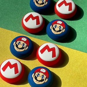May include: Eight colorful silicone controller caps for a gaming console.  Four caps feature a red 'M' on a white background and four caps feature a cartoon image of Mario on a blue background.