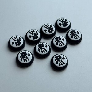 May include: A collection of ten black and white circular decorative buttons. Each button has a white center with a black abstract design and a black border. The buttons are scattered on a light surface.