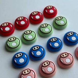 May include: A collection of colorful, mushroom-shaped decorative buttons. The buttons are red, green, blue, and pink, each with a white and brown design. The buttons are arranged on a white surface.