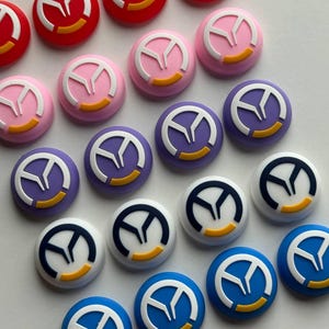 May include: A collection of colorful, round buttons with a stylized emblem. The buttons are in various colors, including red, pink, purple, white, and blue. Each button features a white emblem with a yellow accent.