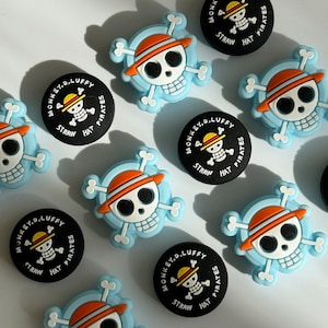 May include: Six blue and white skull and crossbones charms with an orange hat on the skull. Each charm has the text "MONKEY.D. LUFFY" and "STRAW HAT PIRATES" around the skull.