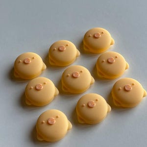 May include: A collection of ten light orange, circular decorative embellishments. Each embellishment features a small, raised pig face with a pink nose and eyes. The embellishments have a smooth, matte finish and are arranged on a light blue surface.