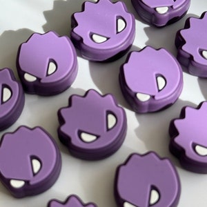 May include: A collection of purple, cartoon-like charms with a jagged edge design. Each charm features a white, D-shaped eye and a white, curved mouth. The charms are arranged on a white surface, with shadows adding depth.