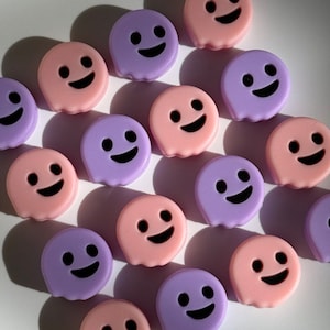 May include: A collection of small, ghost-shaped objects in pastel pink and purple. Each ghost has a smiling face with black eyes and a curved mouth. The objects are arranged on a white surface, casting shadows.