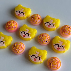 May include: A set of ten yellow and pink resin charms featuring a cartoon character with blonde hair and a pink star. The charms are ideal for adding a touch of whimsy to jewellery, keyrings, or other crafts.