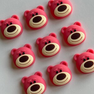 May include: Nine pink bear face shaped buttons with brown eyes and a black nose. The buttons are approximately 1 inch in diameter.