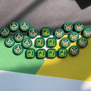 May include: A collection of 20 green and yellow circular game pieces with various symbols. The pieces are arranged in a circle on a gray, green, and yellow surface.