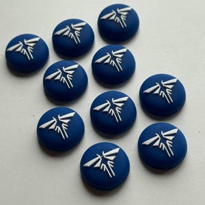 May include: A collection of ten blue, circular buttons with a white graphic design. The design features a stylized, symmetrical shape resembling a flame or insect. The buttons are arranged on a light-colored surface.