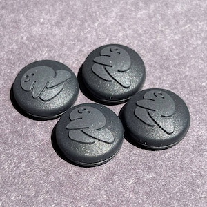 May include: Four black round buttons with a raised design of a cartoon character. The character is a simple, stylized figure with a round head and a body that resembles a blob. The character is depicted in a relaxed, sitting pose with its arms crossed.