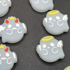 May include: A set of eight small, white, ghost-shaped silicone charms. Some of the ghosts have yellow halos and wings, while others have red horns and a yellow crown.