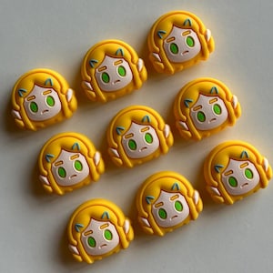 May include: Nine yellow cartoon character charms with a female face and green eyes. The charms are made of a soft, pliable material.