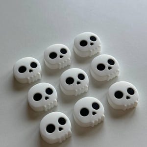 May include: Ten white plastic skull buttons with black eyes. The skulls have a simple design with a rounded shape and a slight drip effect.