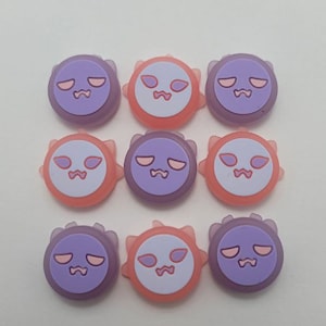 May include: Nine round, cartoon-faced buttons in a grid pattern. The buttons alternate between purple and orange, each with a white face, pink mouth, and D-shaped eyes. The buttons have a spiky border.
