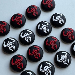 May include: A collection of black, round buttons with a tribal bull design. Some buttons feature a red bull design, while others have a white bull design. The buttons are arranged on a light gray surface.