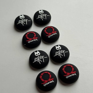 May include: A collection of black, circular controller thumb grips. Some grips feature the word "STRAY" in white with a cat skull logo, while others display a red omega symbol and the words "GOD OF WAR".