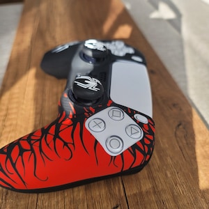 May include: A black, red, and white video game controller with a unique design. The controller features a red base with black vein-like patterns, white buttons, and black thumbsticks with a spider emblem. The controller is resting on a wooden surface.
