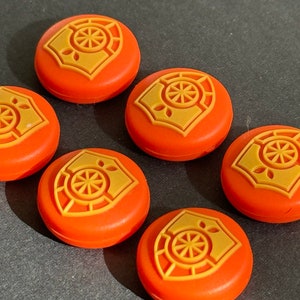 May include: Orange, circular buttons with a gold emblem featuring a stylized wheel design. The buttons are arranged on a dark gray surface. The emblem is raised and has a detailed, geometric pattern. The buttons are likely decorative or functional.