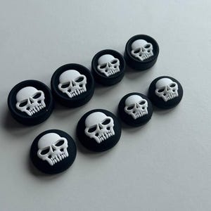 May include: A collection of black and white skull-shaped decorative buttons. The buttons feature a white skull design on a black circular base. The buttons vary in size and are arranged on a light gray surface.