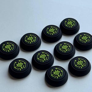 May include: A collection of black controller thumb grips with a neon green design. The grips are round and feature a stylized eye symbol. The image shows multiple grips, suggesting a set or bulk purchase.