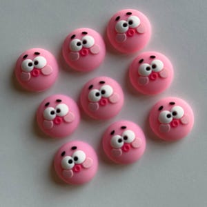 May include: Ten pink, round buttons with cartoon faces. Each button has white eyes, black eyebrows, and pink cheeks and lips. The buttons are arranged on a white surface.