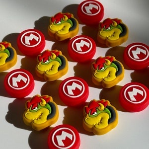 May include: A set of ten red and white controller caps with a white "M" in the center and five yellow and green controller caps with a cartoon image of Bowser, a character from the Super Mario Bros. video game series.