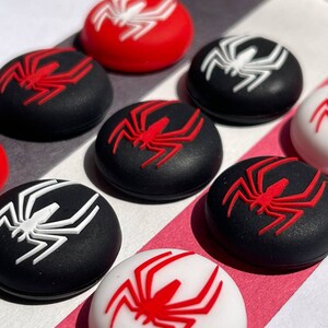 May include: Eight round, black and white silicone controller caps with a red spider design. The caps are arranged in two rows of four.