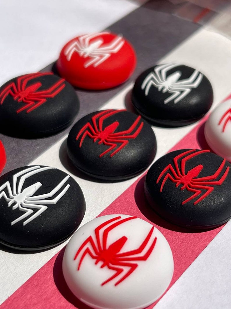 May include: Eight round, black and white silicone controller caps with a red spider design. The caps are arranged in two rows of four.