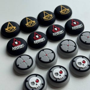 May include: A collection of black and gray controller thumb grips. Some grips feature a gold emblem, others have the text "ASSASSINS CREED" with a red emblem, and others have a gray crosshair or a skull with a red eye.