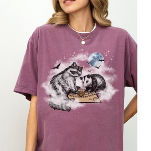 K&ouml;nnte beinhalten: Ein lila T-Shirt mit einem Aufdruck eines Waschb&auml;ren und eines Opossums, die auf einer Wolke sitzen, mit einem Ouija-Brett vor ihnen. Der Aufdruck ist in Schwarzwei&szlig; und der Hintergrund ist dunkelviolett.