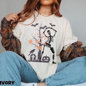 May include: A cream colored t-shirt with a black skeleton dancing in front of a tree with orange leaves. A black crow sits on a tombstone with a white cross. A jack-o-lantern sits at the base of the tree. The text "IVORY" is at the bottom of the image.