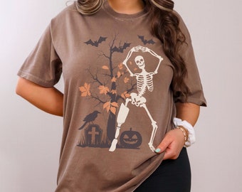 Peut inclure: Un t-shirt marron avec un imprimé d'un squelette dansant devant un arbre aux feuilles orange. Le squelette tient une citrouille et il y a un corbeau sur une tombe en arrière-plan.