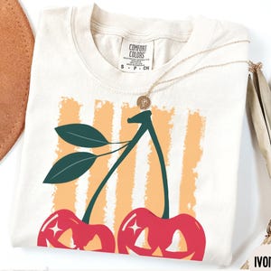 May include: Ivory-colored t-shirt featuring a cherry design. The graphic shows two red cherries with green stems and leaves, set against a background of orange stripes. The shirt has a Comfort Colors tag.