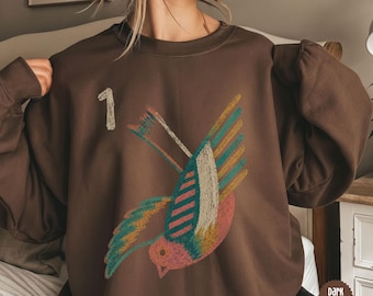 Peut inclure: Un sweat-shirt à col rond marron foncé orné d'un motif d'oiseau en vol, réalisé dans un style texturé rappelant le crayon avec des tons bleu sarcelle, rose et jaune. Le chiffre 1 est imprimé en blanc, façon craie, au-dessus de l'oiseau.