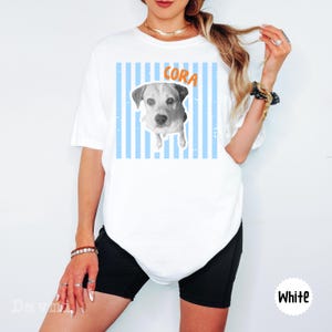 Peut inclure: Un t-shirt blanc &agrave; manches courtes avec un graphique en noir et blanc repr&eacute;sentant un chien, centr&eacute; sur des rayures verticales bleu clair. Le nom CORA est imprim&eacute; en lettres orange au-dessus du chien. Le t-shirt est port&eacute; avec un short noir.