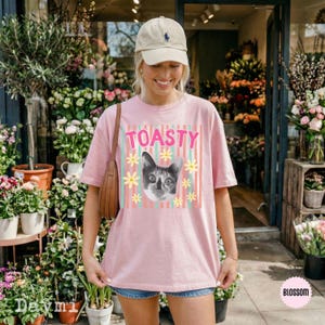 Peut inclure: Un t-shirt rose clair &agrave; manches courtes orn&eacute; d'un motif repr&eacute;sentant une t&ecirc;te de chat entour&eacute;e de marguerites jaunes et du mot TOASTY en lettres roses, sur un fond de rayures verticales bleu sarcelle et roses. Le t-shirt a un col rond.