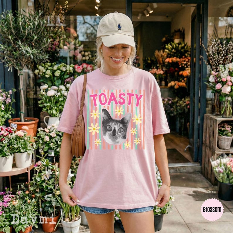 Peut inclure: Un t-shirt rose clair &agrave; manches courtes orn&eacute; d'un motif repr&eacute;sentant une t&ecirc;te de chat entour&eacute;e de marguerites jaunes et du mot TOASTY en lettres roses, sur un fond de rayures verticales bleu sarcelle et roses. Le t-shirt a un col rond.