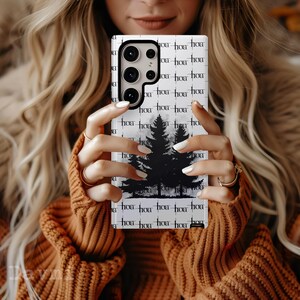 May include: A white phone case featuring a black silhouette of pine trees against a misty background. The case has a repeating pattern of the word "hoa" in a stylized font. The phone case is held by a person wearing an orange sweater.