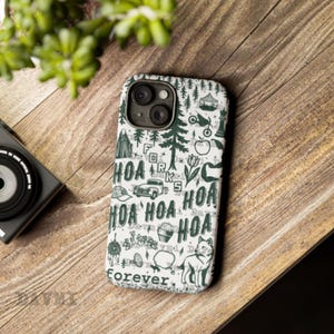 May include: A white phone case with a green and black design featuring the text "HOA" repeated, along with illustrations of trees, a car, and a dog. The case is on a wooden surface, with a camera and greenery in the background.