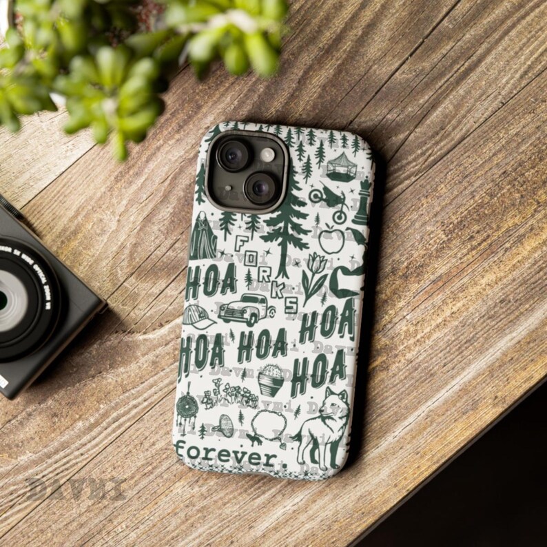 May include: A white phone case with a green and black design featuring the text "HOA" repeated, along with illustrations of trees, a car, and a dog. The case is on a wooden surface, with a camera and greenery in the background.